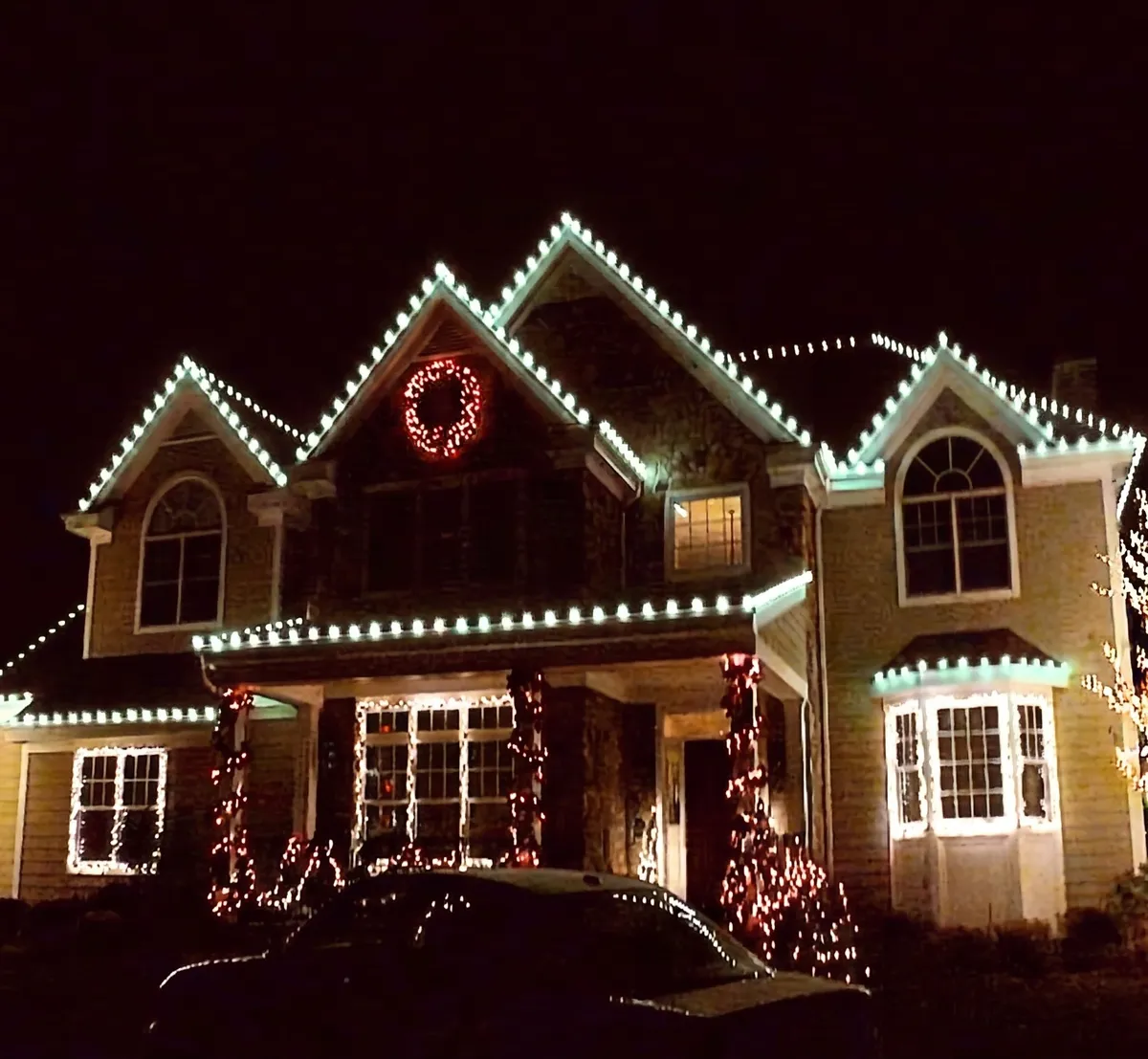 Two-story home with warm white lights tracing every gable and a lit wreath in the center